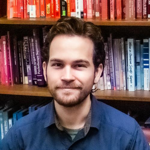 Nico Nelson staff photo; young, white male with brown hair and a short cropped beard; he wears a dark blue shirt and sits in front of a shelf of multicolor books. 