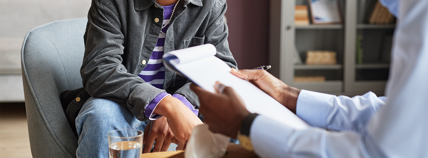 Youth in a grey jacket sits in a char by his therapist, who holds a clipboard. 