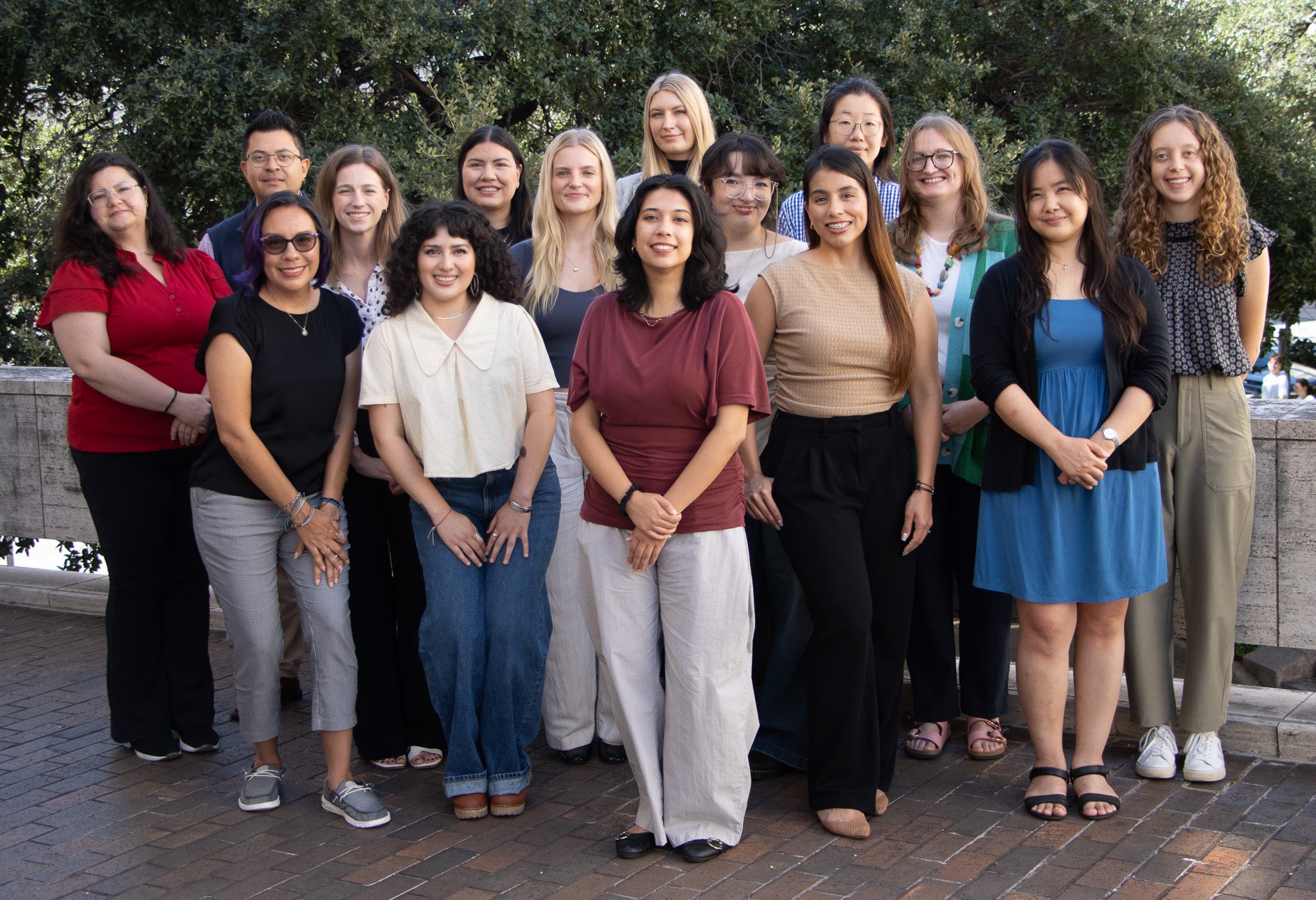 Group photo of the ACT LEND cohort, a group of 15 people standing together outdoors on a brick plaza in front of a large tree and stone wall. The group is arranged in two rows, smiling at the camera.