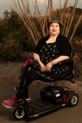 Naomi Ortiz, a light-skinned mestize, sits in their scooter in front of an ocotillo in the desert. They are wearing a fedora hat, hoop earrings, black dress with cacti print and pink boots.  Photo Credit: Jade Beall