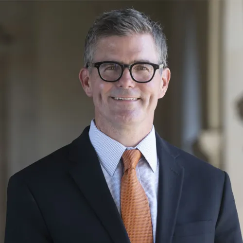 Dean Allan Cole; White adult male, salt and pepper hair, black glasses, black suit with white oxford and burnt orange necktie 