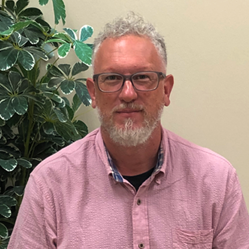 Daniel Meyers headshot: White hair and beard, dark grey glasses; he wears a pink shirt and sits in front of an artificial tree. 