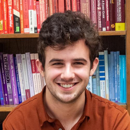 Profile Picture of Alternative Financing Coordinator Cole Glosser; young man with wavy brown hair, dark eyes, smiling; he wears a burnt orange polo and sits in front of a color coordinated bookcase