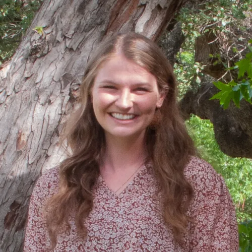 Christiana is a young woman with strawberry-blonde hair. She wears a red and white floral printed blouse and stands in front of a tree. 