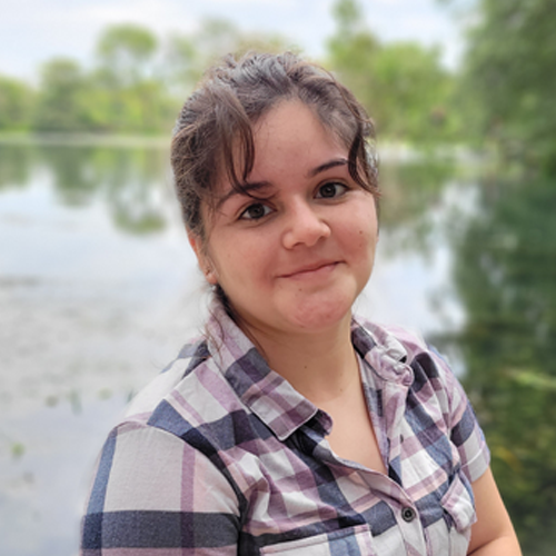 Cristal is a young woman with brown hair tied back. She wears a plaid shirt and sits in front of a lake with trees in the distance. 