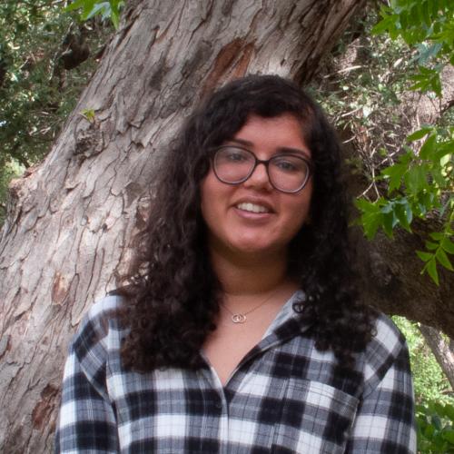 Heena is a young woman with long, curly brown hair and round glasses. She wears a plaid white and black shirt. 