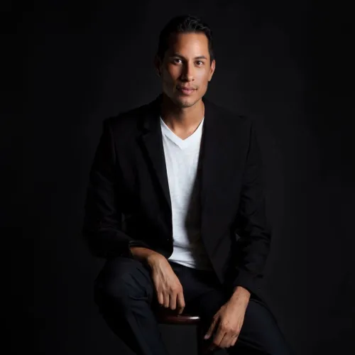 Description: Carlos is a young, Latino male. He is clean shaven with black hair; he wears dark jeans, a black blazer, and a white v-neck shirt; he sits on a stool before a black background.