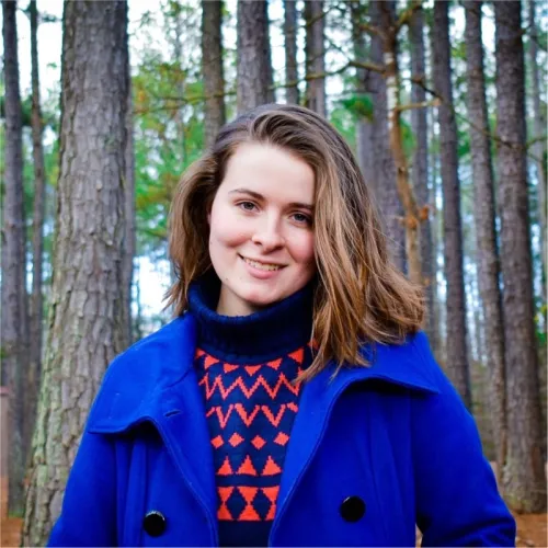 Katheryn is a young woman with brown hair. She wears a blue overcoat and a blue/red pattered sweater. She stands in front of trees in a forest. 
