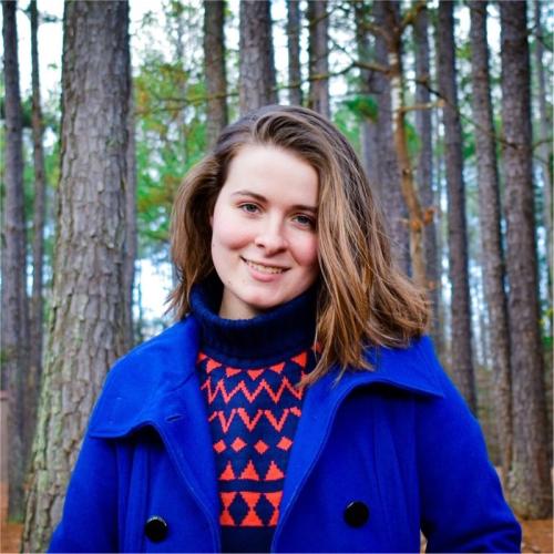 Katheryn is a young woman with brown hair. She wears a blue overcoat and a blue/red pattered sweater. She stands in front of trees in a forest. 