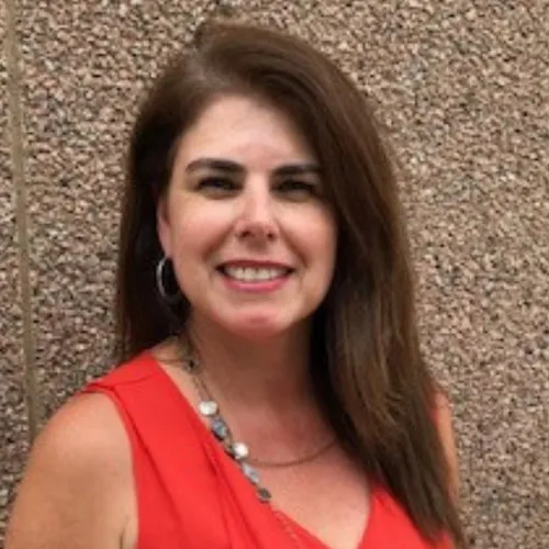 Description: Laura has long brown hair, dark eyes and hoop earrings. She wears a sleeveless red blouse and stands in front of a stone wall. 