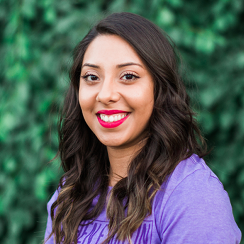 Stephany Brown headshot; Stephany is a young, Latina woman with wavy brown hair. She wears a purple shirt and a red lip. She stands in front of a hedge wall. 