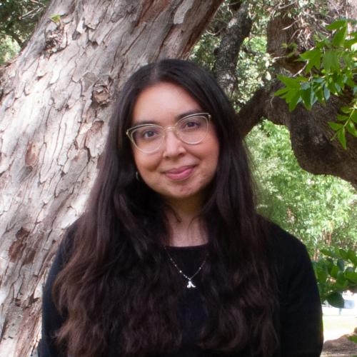 Annabel Garza has long black hair and glasses. She wears a black blouse and stands in front of a tree. 