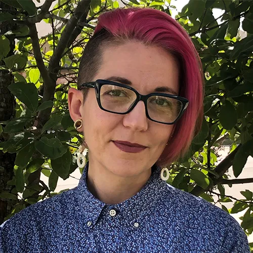 Nick Winges-Yanez Profile picture of Disability Studies Coordinator Nick Winges-Yanez. She has magenta colored hair, black glasses, dark lipstick and wears a blue, floral patterned shirt.