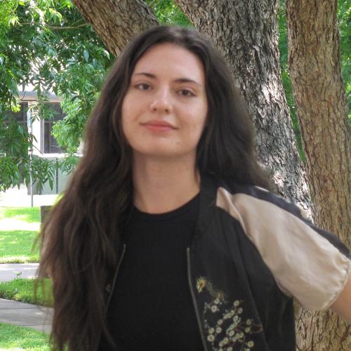 Mary is a tall, white young woman long black hair. She wears a black shirt and a black and white short sleeved, zippered jacket with embroidery on the front. She stand in front of trees.