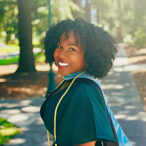 Alexis has short, curly hair. She wears a cap and gown; there is a sidewalk and trees behind her in the distance.