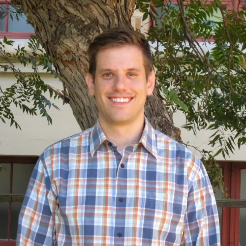 Brad is a young man with dark blonde hair; he wears a blue and orange plaid shirt and stands in front of a tree. 