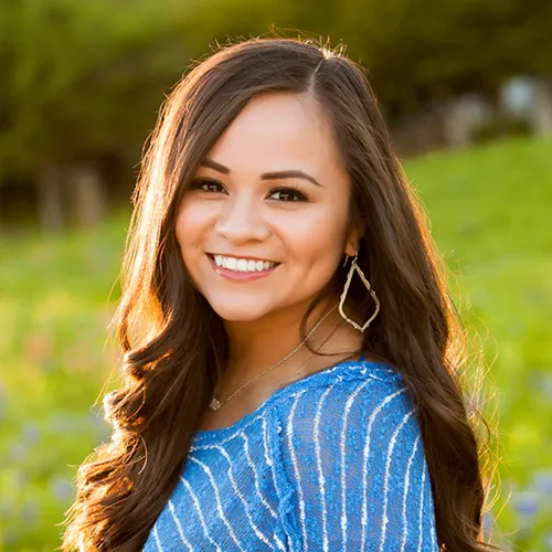 Janie has long brown hair. She wears a blue striped blouse and gold earrings. 