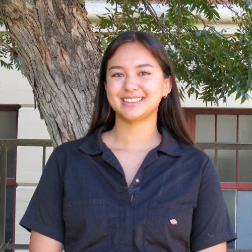 Kaelin has long, black hair. She wears a short-sleeved black jumpsuit and stands in front of a tree.