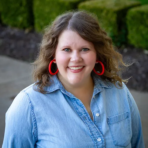 Kari has brown, shoulder-length hair. She wears red hoop earrings and a light denim shirt.