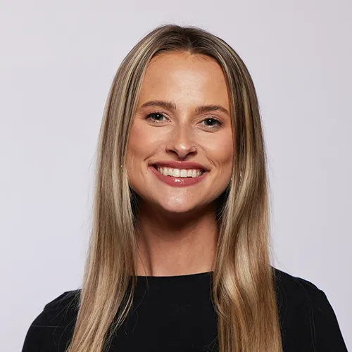 Kayla has long blonde hair. She wears a black dress and stands in front of a purple/grey backdrop.