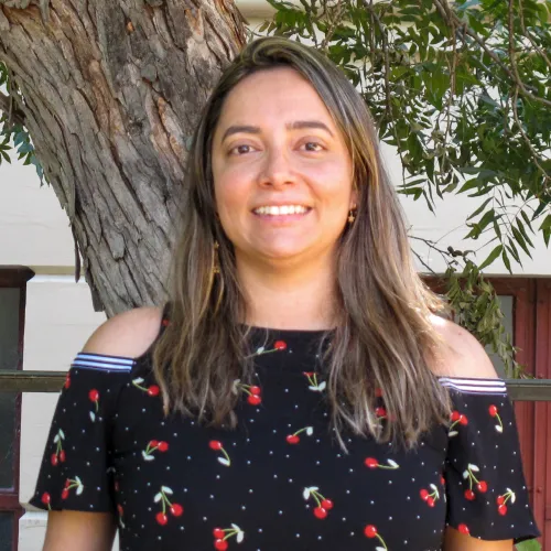 Monique has shoulder length brown hair. She wears a dark blouse with a pattern of cherries. She stands in front of a tree, smiling.