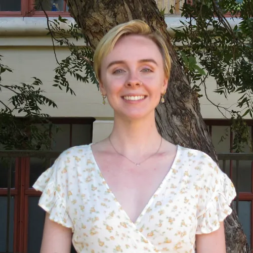 Sara has short blonde hair. She wears a white, floral patterned dress and stands in front of a tree. 