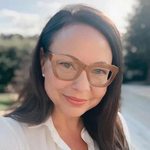 Leslie Jackson has shoulder length, dark brown hair. She wears brown glasses and a white blouse in front of trees.