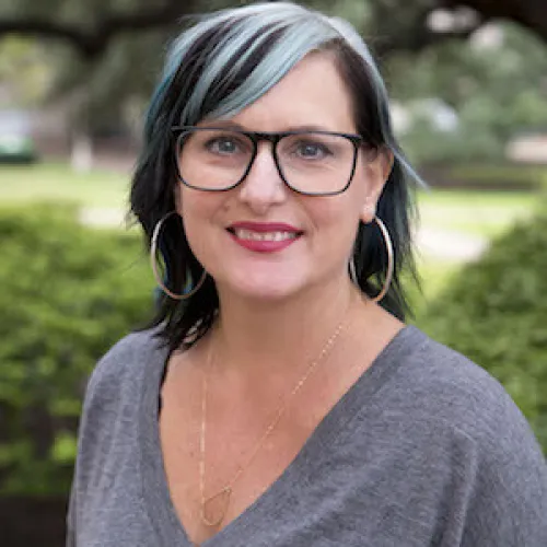 Catherine has striped grey and black hair, hoop earrings and dark eye glasses. She wears a grey blouse and stands in front of greenery.