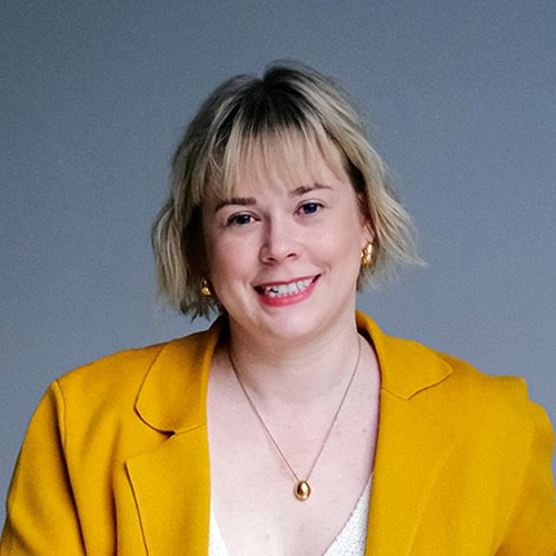 Elizabeth Slade has short blonde hair. She wears a mustard color blazer with a thin, gold necklace. 