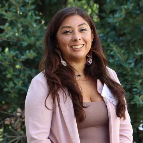 Alyssa is a young Latina woman with long brown hair. She wears a pink blazer and a purple top underneath.