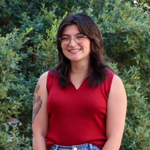 Tais is a young woman with shoulder length brown hair, glasses, and a red, sleeveless top.