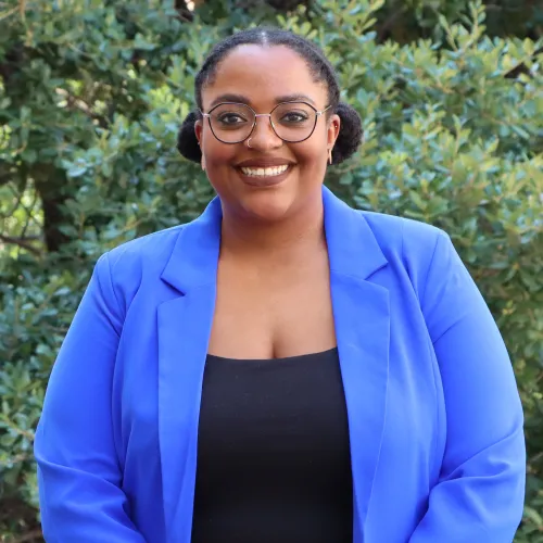 Jerrica has dark hair, pulled into two buns. She wears round glasses, a light blue blazer and a black top underneath.