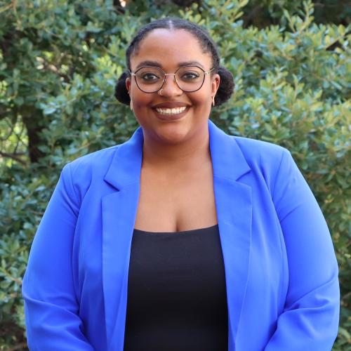 Jerrica has dark hair, pulled into two buns. She wears round glasses, a light blue blazer and a black top underneath.
