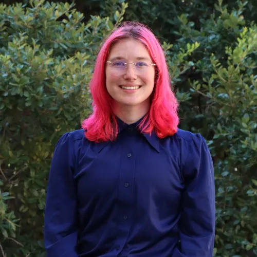 Ari is a young person with fair skin. They have vibrant pink hair, thin glasses and a navy blue blouse.