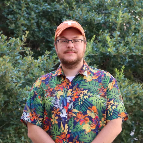 Benny has reddish brown hair and beard. He wears a vibrant Hawaiian shirt and an orange UT cap.