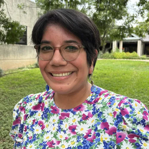 Delyla headshot - short brown hair with a blue/purple floral shirt and glasses.
