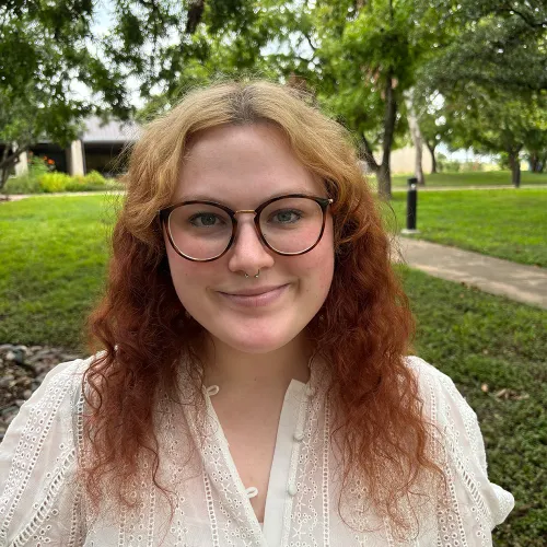 Adra has red hair with blonde roots. She has large, round glasses and wears a lacey white blouse.