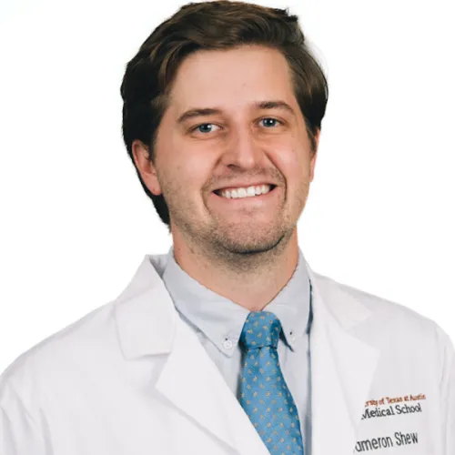 Cameron has short brown hair and a cropped beard. He wears a white doctor's lab coat over a light blue shirt with a blue, polkadot necktie.