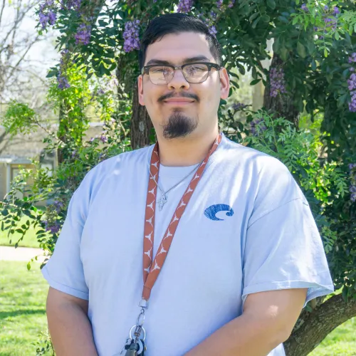 Alex has short black hair with black-rimmed glasses and a goatee. He wears a light blue t-shirt with a burnt orange UT Lanyard and a silver necklace.