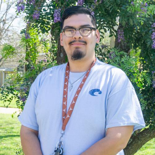 Alex has short black hair with black-rimmed glasses and a goatee. He wears a light blue t-shirt with a burnt orange UT Lanyard and a silver necklace. 