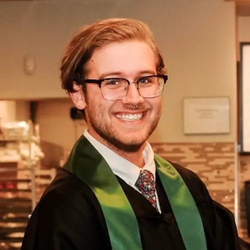 Dalton has strawberry blond hair, dark framed glasses and a short cropped beard. He wears a black graduation gown with a green sash.