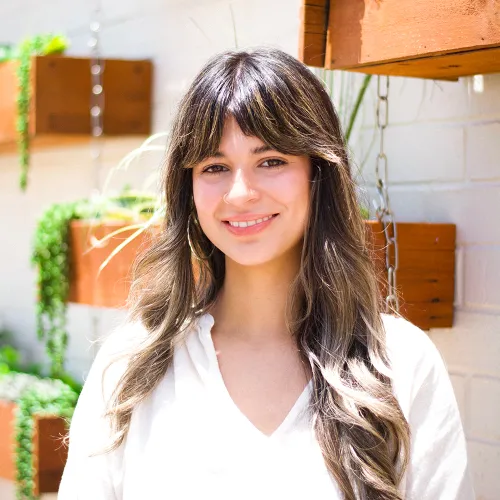 Destiny has long, wavy brown hair with blonde highlights. She wears gold hoop earings and a white, v-neck blouse. She stands in front of a white brick wall with hanging wooden planters.