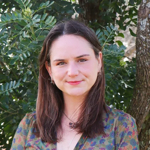 Image of Michelle Phillips; Michelle has long brown hair and wears a multi-colored blouse in front of green foliage.