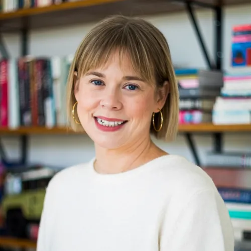 Elizabeth Slade has dark blonde hair, gold hoop earrings, and a dark red lip. She wears a white blouse and stands in front of a bookshelf with multi-colored books.