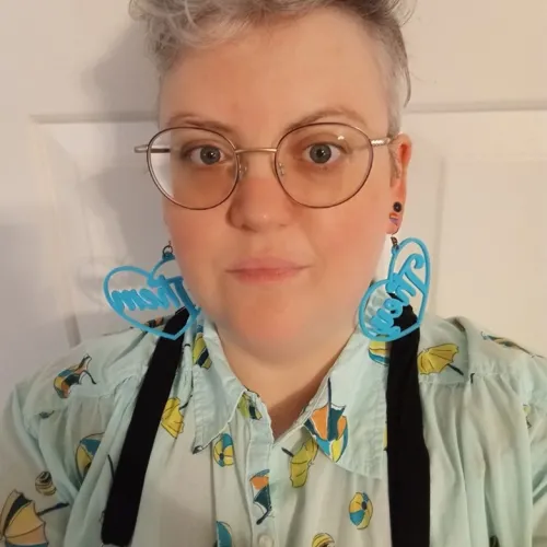 A person with short curly hair wearing round glasses, blue heart shaped earrings, and a patterned shirt.