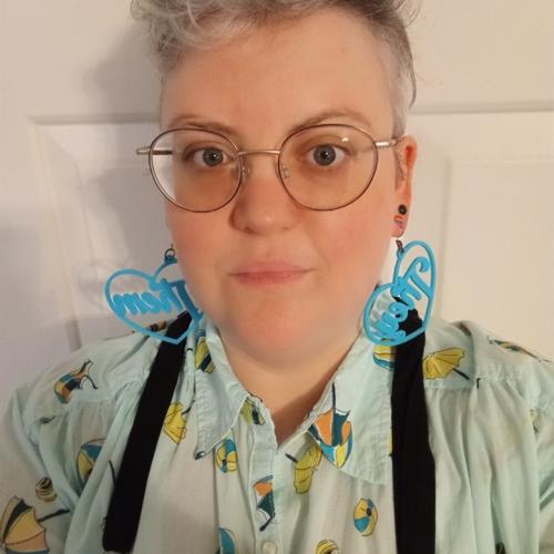 A person with short curly hair wearing round glasses, blue heart shaped earrings, and a patterned shirt. 