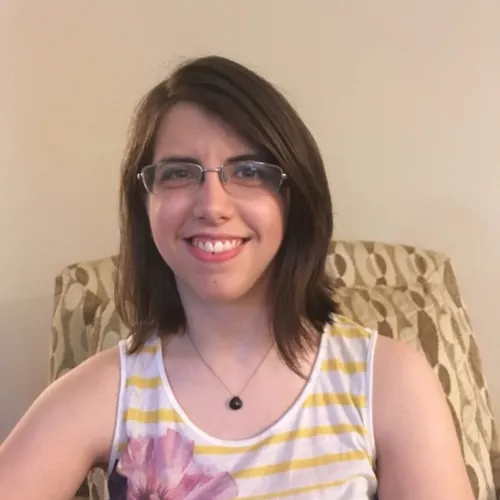 A person with medium length hair smiling wearing glasses, a necklace and a tank top with yellow stripes on the top and a purple flower.