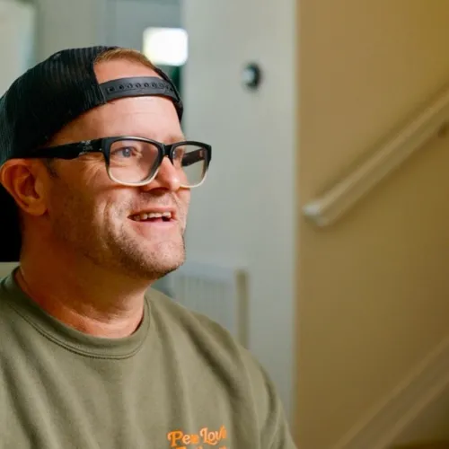 A person to the side, smiling with a hat, wearing glasses and a shirt. 