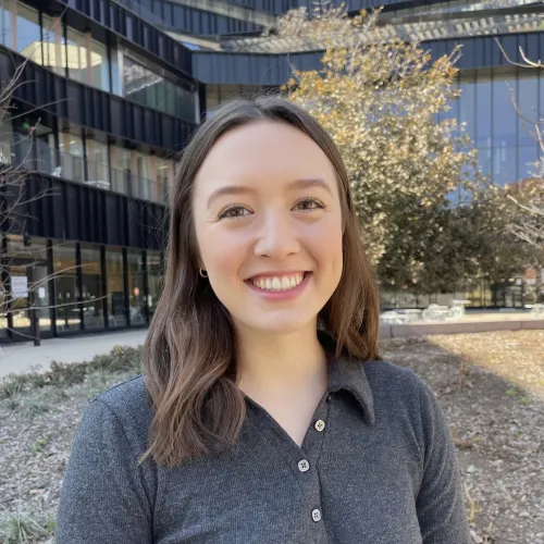 Libby Banks has brown hair and wears a dark grey long-sleeve polo. She stands in front of a modern building.