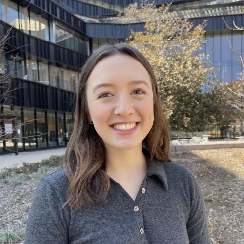 Libby Banks has brown hair and wears a dark grey long-sleeve polo. She stands in front of a modern building.